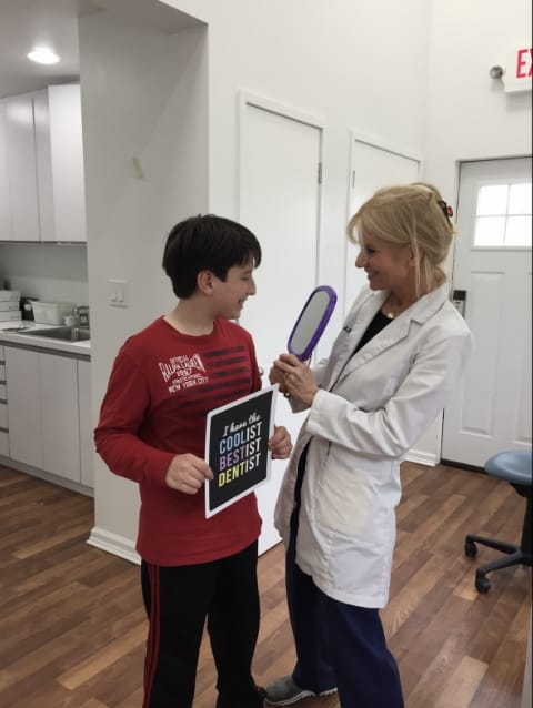 Dr. Danoff holding mirror to boys face. Boy is holding sign that says I have the collist bestist dentist