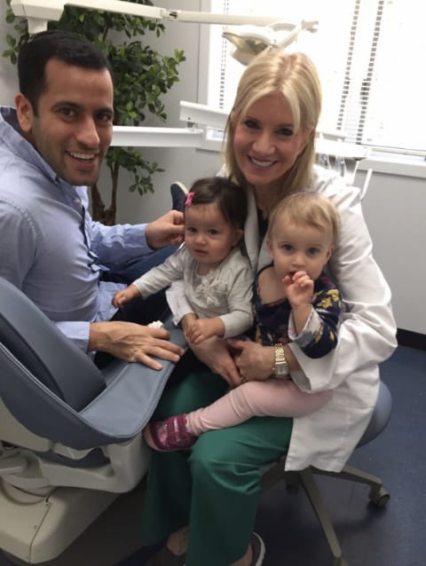 Dr. Danoff holding two babies while sitting next to dental chair with man on it